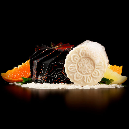 Decorative foaming citrus shampoo bar with floral pattern beside black and orange packaging, surrounded by orange and lemon slices and rice