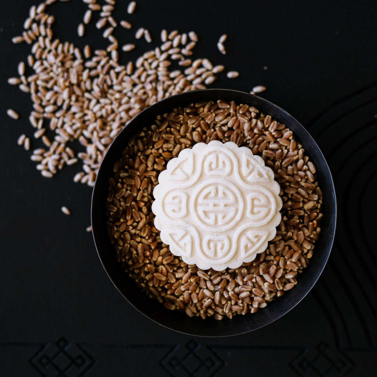 Decorative white mooncake with intricate pattern on a bowl of wheat grains on black background