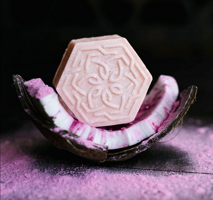 Pink hexagonal moisturizing shampoo bar with intricate design on purple powder and coconut shell